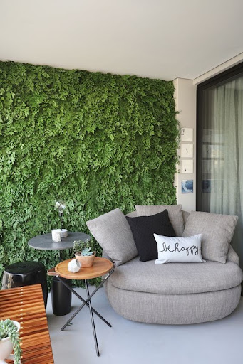 Varanda de apartamento com puff grande para área externa, mesinhas laterais de apoio e parede com jardim vertical de plantas permanentes 
