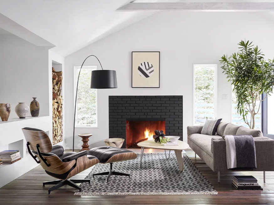 Na foto uma sala de estar com paredes brancas, lareira preta, luminária de chão, sofá cinza e poltrona de design assinado Eames Lounge Chair