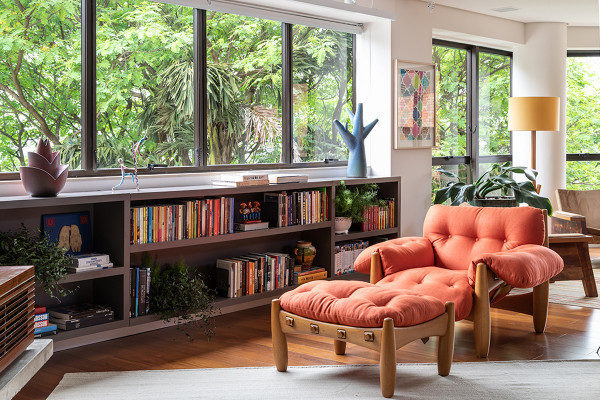Na foto sala de estar com grande janela, embaixo, um móvel com diversos livros e a frnete uma poltrona com design assina na cor laranja.