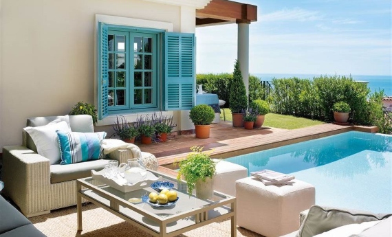 Área da piscina de uma casa decorada com sofá e puffs brancos, mesa central de vidro e plantas 